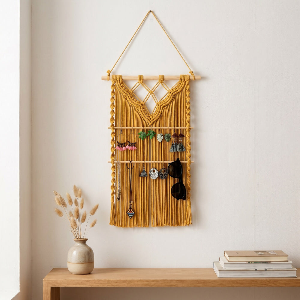 A mustard yellow macramé wall hanging with wooden bars for organizing jewelry, displayed on a white wall above a table.